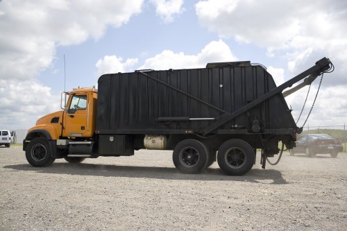 A large industrial waste collection truck parked on a gravel surface, featuring a yellow cab with a sturdy black waste container attached at the rear. The container has a textured, matte finish and is fitted with a side-loading mechanism, including a long horizontal arm extending from the back to facilitate waste collection. The truck's black wheels are visible beneath the container, and the environment appears to be an outdoor storage or loading area with a partly cloudy sky overhead. In the background, other vehicles or equipment can be seen, indicating a commercial waste management setting, possibly in the Waltham Forest area. The scene is well-lit with natural daylight, emphasizing the utilitarian design of the vehicle, which is used for rubbish removal and waste collection services by companies like Commercial Waste Waltham Forest.