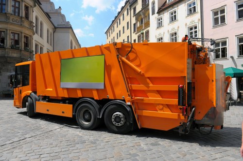 A large, bright orange rubbish collection vehicle parked on a cobblestone street in an urban area with classic European-style buildings in the background. The vehicle is a modern waste disposal truck with a smooth, glossy exterior, featuring a reflective green panel on its side. It has large black rubber tires with silver rims and is equipped with various mechanical elements, including hydraulic hoses and rear loading mechanisms. The scene is set outdoors during daylight, with a partly cloudy sky visible above the rooftops. The surroundings suggest a busy town or central Waltham Forest location, accommodating waste collection services provided by Commercial Waste Waltham Forest, focusing on rubbish removal for local businesses or residents in the area.