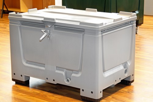 A large, modern plastic storage container with a light grey finish, situated indoors on a polished wooden floor. The container features a secure lid with integrated handles on the sides and appears to be used for storing or transporting waste or materials. Its surface has a smooth, slightly textured appearance, with visible seams and reinforced edges for durability. The container is positioned centrally in the image, with part of a green waste bin and other objects visible in the background, indicating a setting associated with waste management or storage. The lighting is bright and even, highlighting the container's clean and sturdy construction, consistent with equipment used in commercial rubbish removal services by companies like Commercial Waste Waltham Forest, serving areas such as Waltham Forest and nearby locations.