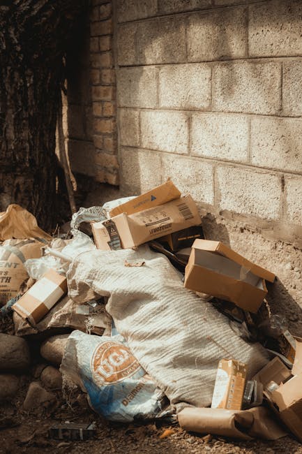 A pile of mixed rubbish accumulated on a gravel surface outdoors, consisting of black and white plastic garbage bags, a yellow plastic container, and an old, dirty armchair with worn fabric and visibl
