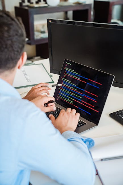 A young man with dark, curly hair, wearing a white t-shirt with maroon and purple horizontal stripes, is seated at a desk in an office environment. He is focused on typing on a black keyboard with a c